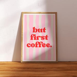 Könnte beinhalten: Ein gerahmter Druck mit dem Text "but first coffee" in Rot, vor einem rosa-cremefarbenen gestreiften Hintergrund. Der Holzrahmen steht auf einer Holzoberfläche und lehnt an einer weißen Wand.