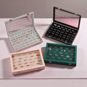 Puede incluir: Cuatro estuches expositores de joyas en varios colores: gris claro, negro, rosa y verde. Cada estuche está abierto, revelando múltiples pares de pendientes. Los estuches tienen tapas transparentes y están colocados sobre una superficie rosa.