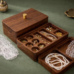 Puede incluir: Una caja de joyería de madera oscura con múltiples compartimentos, incluyendo ranuras para anillos y secciones para collares y pulseras. La caja está abierta, revelando varias joyas. La tapa tiene un botón dorado.