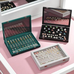 Może przedstawiać: Trzy gabloty na biżuterię ze szklanymi pokrywami. Jedna jest zielona, ​​jedna czarna, a druga szara. Każda gablota jest wypełniona pierścionkami o różnych wzorach i kolorach, osadzonymi na aksamitnym wnętrzu. Gabloty są otwarte, prezentując biżuterię.