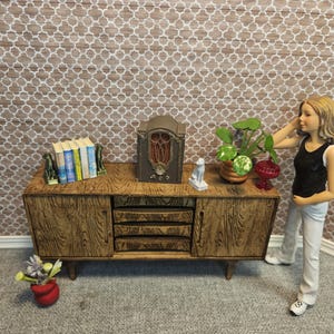 May include: A miniature wooden mid-century modern style cabinet with a radio, books, and a plant. The cabinet has a wood grain finish and is set against a patterned background. A figurine of a woman is standing next to the cabinet.