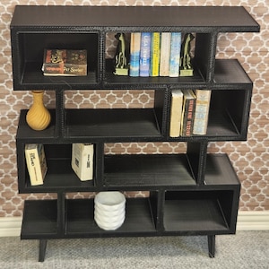 May include: Black miniature bookshelf with multiple cubby spaces. The bookshelf holds miniature books, a yellow vase, and a white decorative item. The books include titles by Stephen King and Lisa Jewell. The bookshelf is on a gray surface.