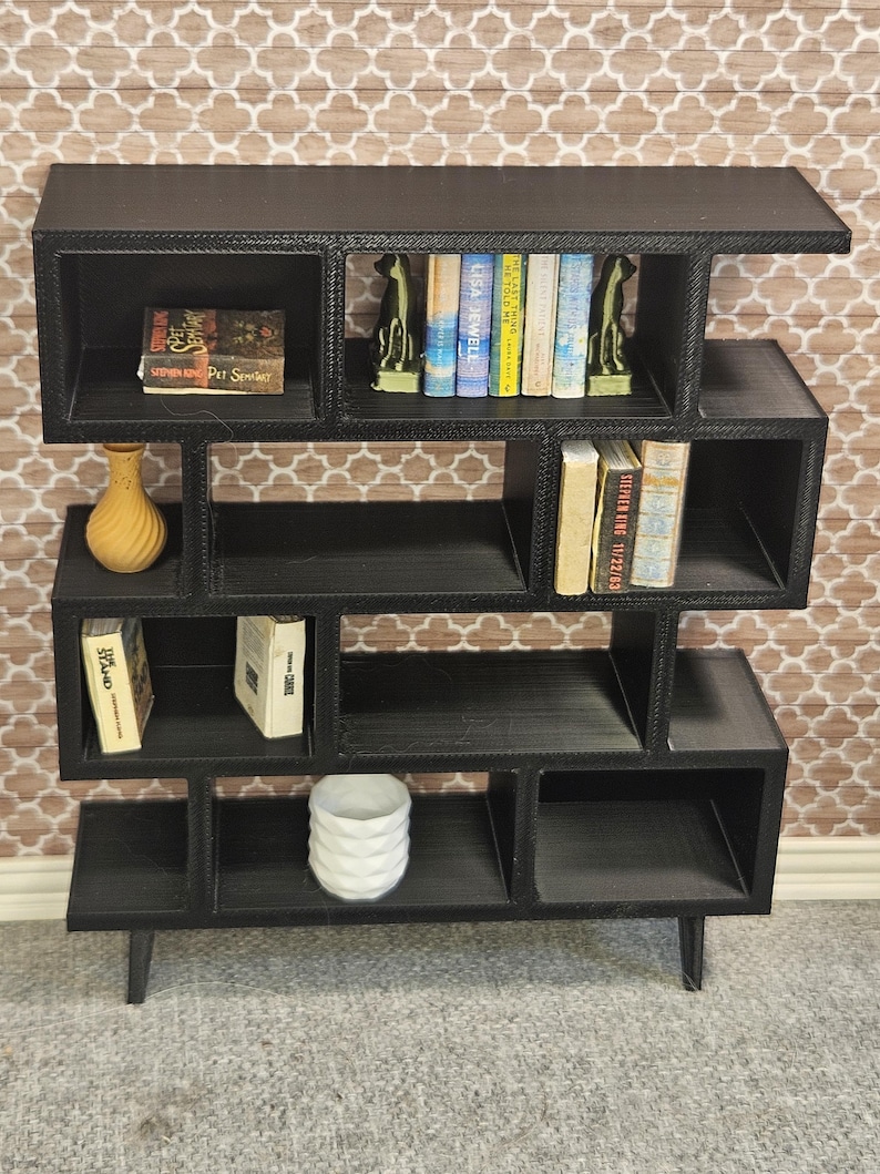 May include: Black miniature bookshelf with multiple cubby spaces. The bookshelf holds miniature books, a yellow vase, and a white decorative item. The books include titles by Stephen King and Lisa Jewell. The bookshelf is on a gray surface.