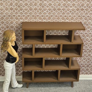 May include: A brown, multi-compartment bookshelf with a mid-century modern design. The bookshelf has a series of open cubbies and shelves of varying sizes. The unit is made of wood and stands on four short legs. The background features a patterned wallpaper.