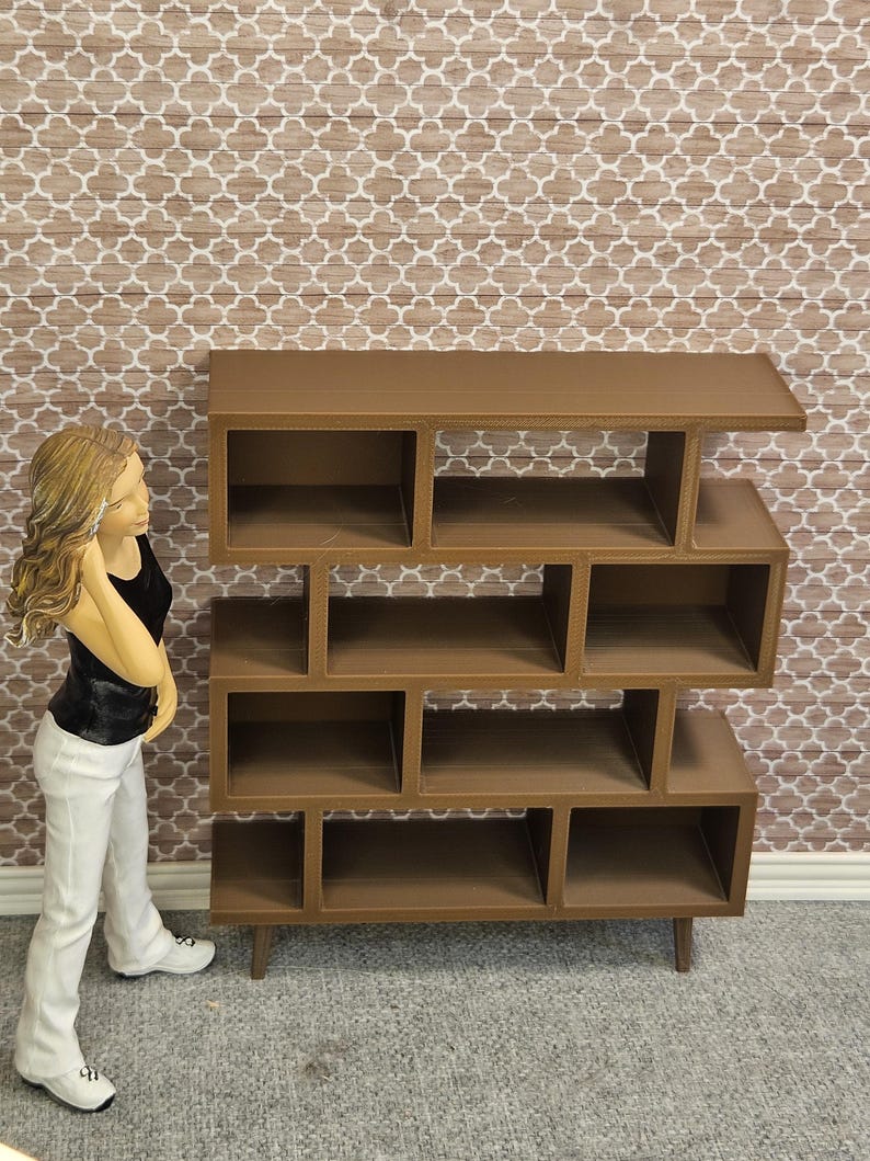 May include: A brown, multi-compartment bookshelf with a mid-century modern design. The bookshelf has a series of open cubbies and shelves of varying sizes. The unit is made of wood and stands on four short legs. The background features a patterned wallpaper.