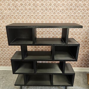 May include: A black, multi-tiered shelf unit with an open, geometric design. The unit features several cubby-like compartments of varying sizes, supported by short legs. The shelf is made of a dark material and is suitable for displaying decorative items.