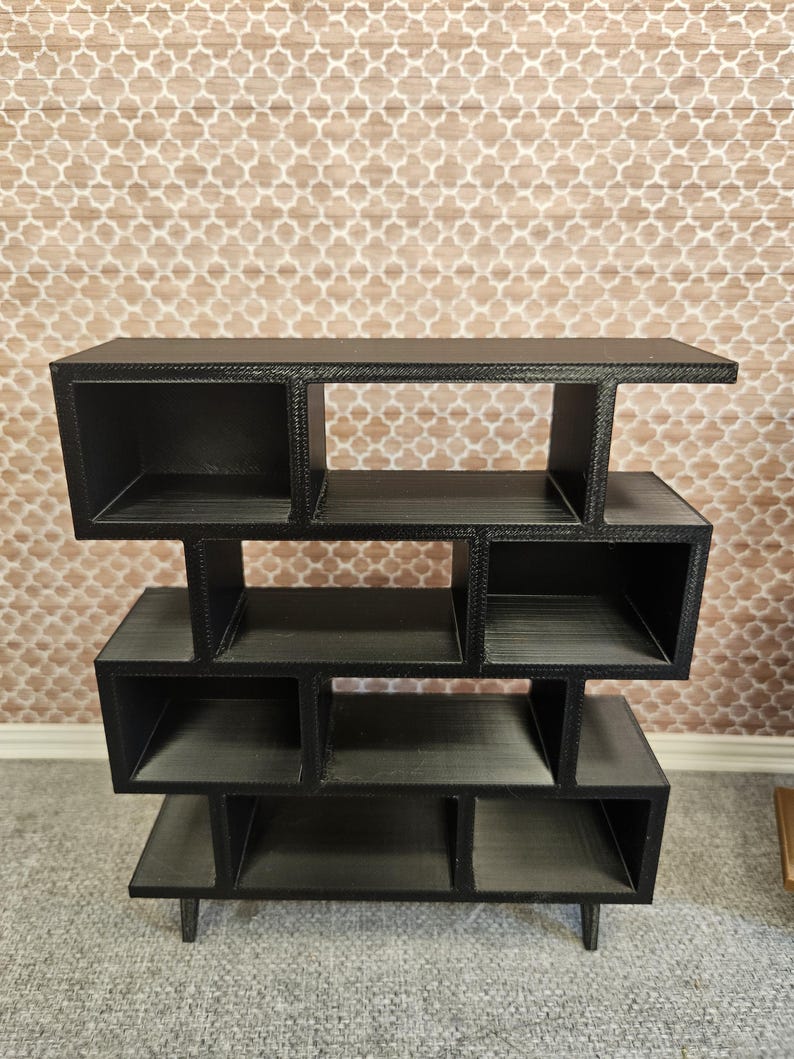 May include: A black, multi-tiered shelf unit with an open, geometric design. The unit features several cubby-like compartments of varying sizes, supported by short legs. The shelf is made of a dark material and is suitable for displaying decorative items.
