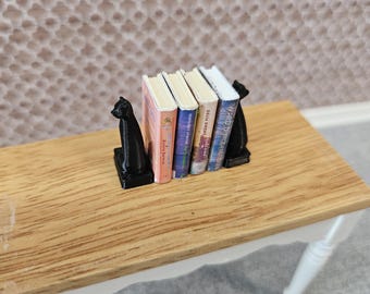 May include: Miniature bookshelf set with four colorful books held by black cat bookends. The books have different colored spines and titles. The set is on a small wooden table with white legs, creating a charming, whimsical scene.
