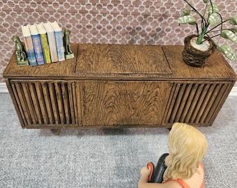 May include: A miniature wooden cabinet with a textured surface, featuring vertical slats on the front. It has a small plant in a woven pot and a stack of books with bookends. A figurine of a person is sitting in front of the cabinet.