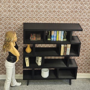 May include: A black, modern dollhouse bookshelf with multiple cubby shelves. The bookshelf is filled with miniature books, a vase, and decorative items. A figurine of a person is standing next to the bookshelf.