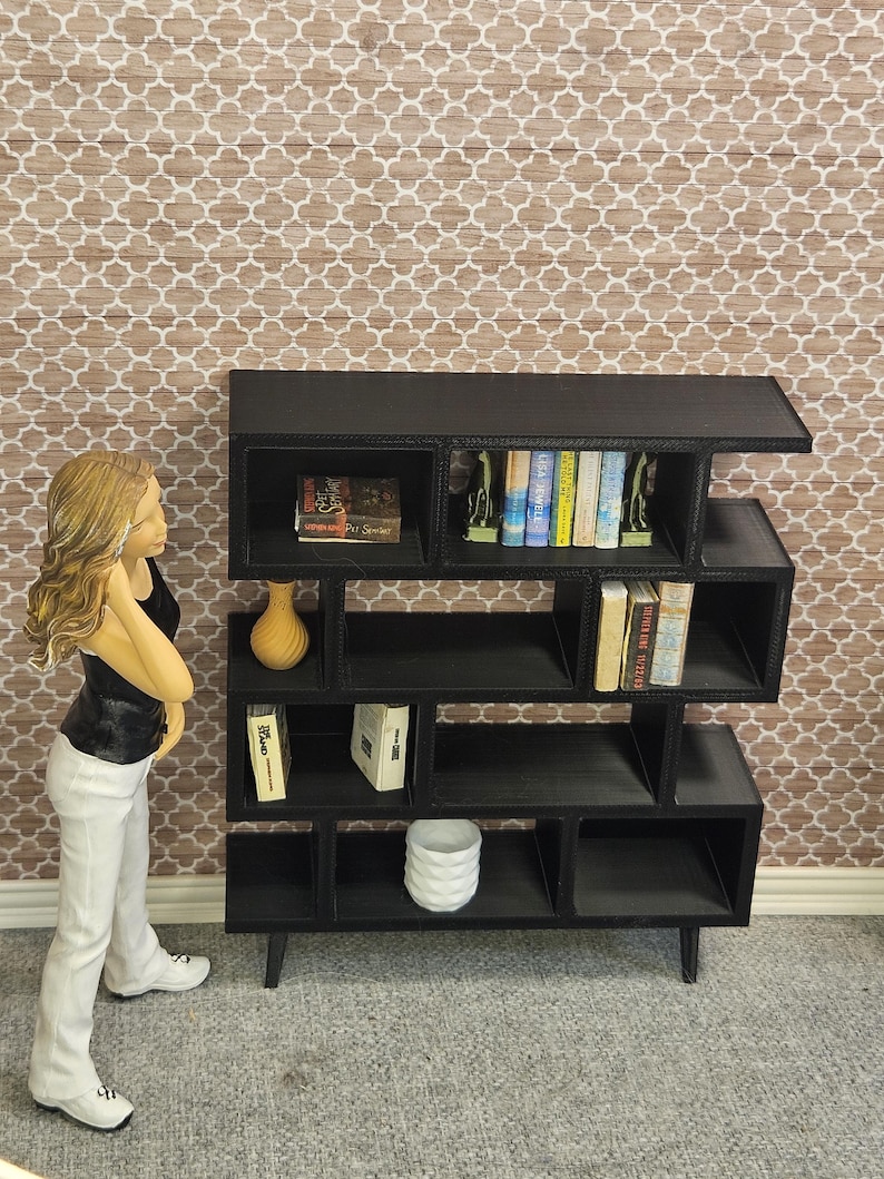 May include: A black, modern dollhouse bookshelf with multiple cubby shelves. The bookshelf is filled with miniature books, a vase, and decorative items. A figurine of a person is standing next to the bookshelf.