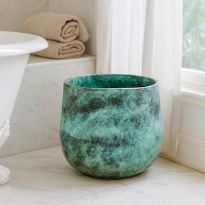 Può includere: Un grande vaso decorativo rotondo di colore turchese con una superficie strutturata. Il vaso presenta un motivo marmorizzato marrone scuro e poggia su una superficie di marmo. Lo sfondo comprende una vasca da bagno bianca, asciugamani arrotolati e una finestra.
