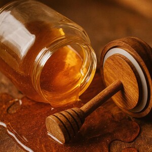 Peut inclure: Un pot de miel en verre, partiellement renversé sur une surface en bois. Une cuillère à miel en bois et un couvercle sont à côté du pot. Le miel est d'une riche couleur ambrée, et le bois a une teinte brun chaud.