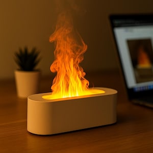 Op de afbeelding: Een witte, rechthoekige lamp met vlameffect straalt helder oranje en geel licht uit, dat lijkt op een flakkerend vuur. De lamp staat op een houten oppervlak, met een laptop en een kleine plant in pot op de achtergrond. Decoratieve woonaccessoire.