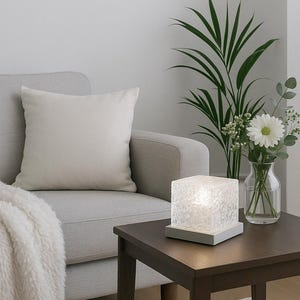 Può includere: Una scena di soggiorno moderno presenta una lampada quadrata in vetro strutturato su un tavolino in legno scuro. Un vaso di fiori bianchi è posto accanto alla lampada. Un divano grigio chiaro con un cuscino color crema e una soffice coperta completano la scena.