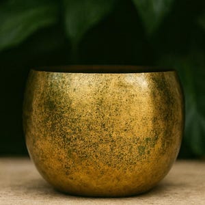 Peut inclure: Un bol doré avec une surface texturée. Le bol a une forme arrondie et un intérieur sombre. L'arrière-plan présente des feuilles vertes floues, créant une esthétique naturelle.