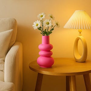 Könnte beinhalten: Ein modernes Wohnzimmer mit einer pinkfarbenen, bauchigen Vase mit weißen Gänseblümchen. Eine gelbe Lampe mit plissiertem Schirm steht auf einem hölzernen Beistelltisch. Ein gerahmtes abstraktes Kunstwerk hängt an der Wand.
