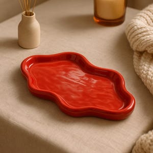 Può includere: Un vassoio in ceramica rosso lucido dalla forma organica e ondulata. Il vassoio è appoggiato su una superficie dai toni neutri, con un piccolo vaso di fiori secchi e una candela accesa in un barattolo di vetro sullo sfondo. È visibile anche una coperta di maglia color crema.