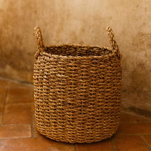 Puede incluir: Una cesta cilíndrica tejida con fibras naturales de color tostado, que presenta una textura gruesa y trenzada en todo el cuerpo y dos asas resistentes y verticales unidas al borde superior para facilitar el transporte y almacenamiento.
