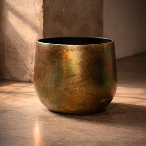 Könnte beinhalten: Ein abgerundeter Pflanztopf mit einer verwitterten, metallischen Oberfläche in gesprenkelten Bronze-, Kupfer- und Grün-Oxidationstönen. Die Außenseite wirkt texturiert und gealtert, während der innere Rand glatt und in mattem Schwarz gehalten ist.
