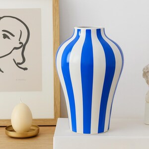 Peut inclure: Un vase en céramique à rayures bleues et blanches. Le vase a une forme arrondie avec un col étroit. Les rayures sont verticales et espacées uniformément. Le vase est posé sur une surface blanche.
