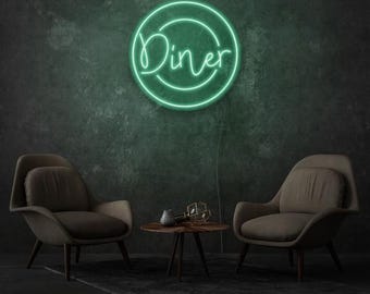 May include: A green neon "Diner" sign in a circular frame is displayed on a dark green wall. Two armchairs and a small table with decorative items are in front of the wall, creating a retro diner theme.