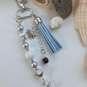 Op de afbeelding: Een zilveren sleutelhanger met een lichtblauwe kwast, een schelpenbedel en een streng witte, grijze en zilveren kralen. De sleutelhanger is versierd met een kleine zwarte kraal en een bloemvormige bedel. Schelpen op de achtergrond.