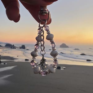 May include: A beaded keychain with a silver clasp and chain. The keychain features a teardrop-shaped design made of white and purple beads, with a small silver charm. The background shows a beach and ocean at sunset.