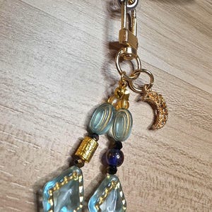 May include: A gold phone charm with a lobster clasp, featuring a beaded chain with blue and gold accents. The chain includes blue diamond-shaped beads, gold cylindrical beads, and a gold crescent moon charm.