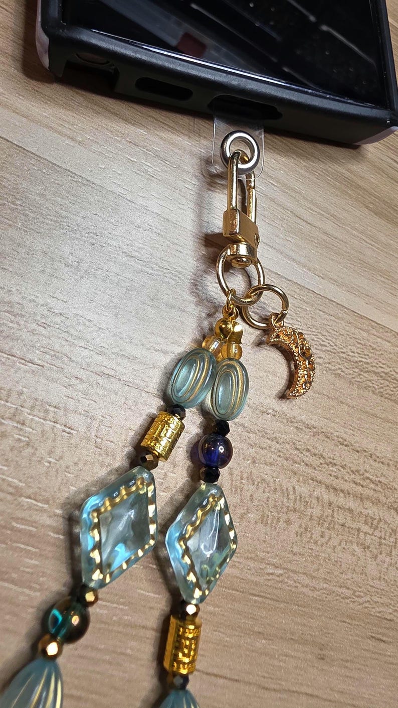 May include: A gold phone charm with a lobster clasp, featuring a beaded chain with blue and gold accents. The chain includes blue diamond-shaped beads, gold cylindrical beads, and a gold crescent moon charm.