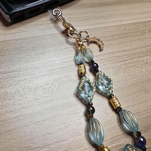 May include: A beaded phone charm with a gold clasp and a crescent moon charm. The charm features light blue, gold, and dark blue beads in various shapes, including diamond and oval. The charm is attached to a black phone case.