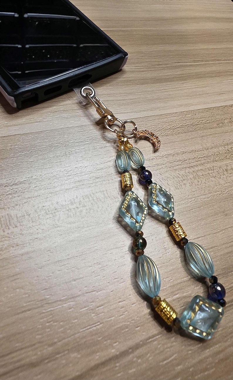 May include: A beaded phone charm with a gold clasp and a crescent moon charm. The charm features light blue, gold, and dark blue beads in various shapes, including diamond and oval. The charm is attached to a black phone case.
