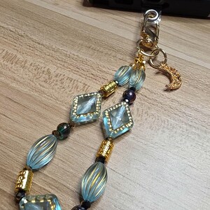 May include: A decorative phone charm with light blue, gold, and dark blue beads. The charm features square and oval beads with gold accents, a gold-coloured clasp, and a small crescent moon charm.