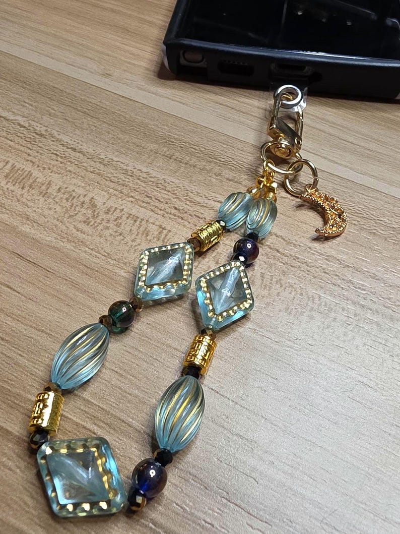 May include: A decorative phone charm with light blue, gold, and dark blue beads. The charm features square and oval beads with gold accents, a gold-coloured clasp, and a small crescent moon charm.