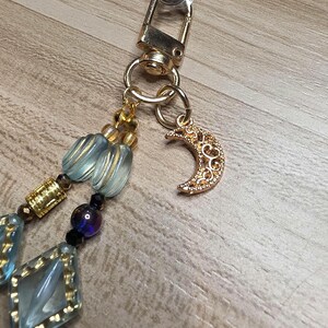 May include: A gold-coloured phone charm with a crescent moon pendant and a beaded strap. The strap features light blue and gold-coloured beads, along with a gold-coloured clasp.