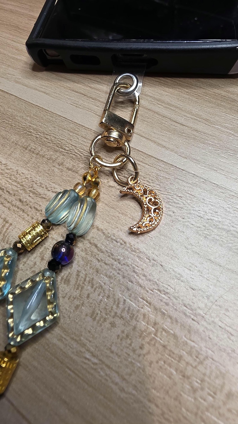 May include: A gold-coloured phone charm with a crescent moon pendant and a beaded strap. The strap features light blue and gold-coloured beads, along with a gold-coloured clasp.
