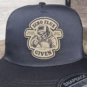 Peut inclure: Une casquette trucker noire avec un écusson beige illustrant un squelette portant un masque de soudage et tenant un chalumeau. Le texte indique ZERO FLUX GIVEN, WELDER BY CHOICE et BUILT BY FIRE. Un autocollant circulaire sur la visière indique SNAPBACK.