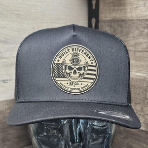 May include: A black snapback cap featuring a circular patch on the front. The patch displays a skull wearing a tactical helmet over a stylised American flag background, with the text BUILT DIFFERENT, RF CO., and MENTALITY, DISCIPLINE, PURPOSE.