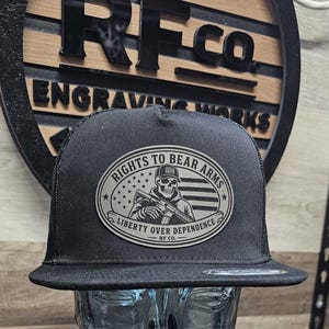 Peut inclure: Une casquette de camionneur noire avec un écusson ovale central sur fond gris. L'écusson représente un crâne portant une casquette, tenant un fusil, avec un drapeau américain. Le texte indique : RIGHTS TO BEAR ARMS, LIBERTY OVER DEPENDENCE, RF CO.