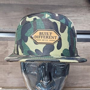 May include: Camouflage print baseball cap with a mesh back. The front of the cap features a wooden patch with the words "BUILT DIFFERENT RF CO." in black lettering. The cap is displayed on a mannequin head.