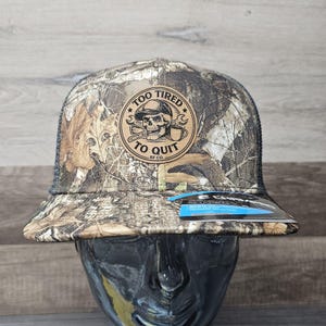 May include: Camouflage print baseball cap with a black mesh back. The cap features a circular patch with a skull wearing a hard hat, wrenches, and the text "TOO TIRED TO QUIT". The RF CO. logo is also visible.