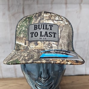 May include: A camouflage-patterned baseball cap with a mesh back. The front features a rectangular patch with the words "BUILT TO LAST" and "RF CO." in black. A blue and white tag is attached to the brim.