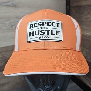 May include: Orange and white trucker hat featuring the text "RESPECT THE HUSTLE" on a white patch. The hat has a curved brim and white mesh panels. The text "RF CO." is below the main text.
