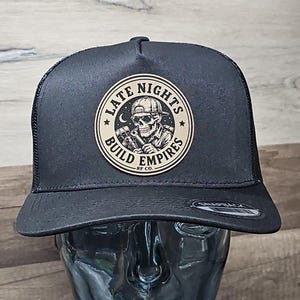 May include: Black trucker hat with a mesh back and a curved peak. The hat displays a circular patch with a skull graphic, the words "LATE NIGHTS BUILD EMPIRES", and a crescent moon and stars.