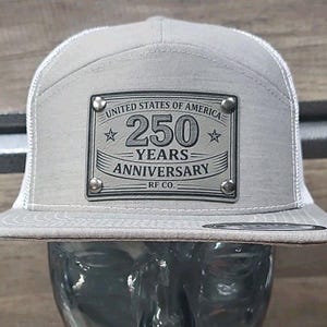 May include: A grey and white trucker cap with a flat peak. The front panel displays a metal plaque with the text "UNITED STATES OF AMERICA 250 YEARS ANNIVERSARY RF CO." The cap has a mesh back.