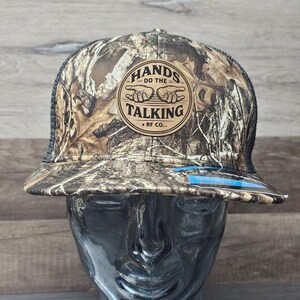 May include: A camouflage print baseball cap with a black mesh back. The cap features a circular patch with the words "HANDS DO THE TALKING" and "RF CO." in a brown and tan colour scheme.