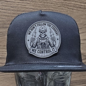 May include: A black trucker hat featuring a circular patch on the front. The patch displays a skull wearing a hard hat, holding lightning bolts, with the text WE DON'T FOLLOW THE CURRENT WE CONTROL IT and RF CO. printed in a black and white design.