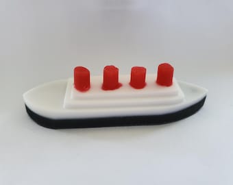 May include: A miniature ship model with a white hull, black base, and four red smokestacks. The model has a layered design, with the smokestacks positioned on a white deck. The overall shape is reminiscent of a classic ocean liner.