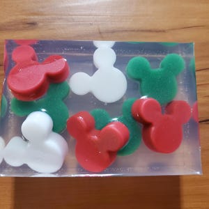May include: A clear rectangular block containing Mickey Mouse-shaped soap pieces in red, white, and green. The soap pieces are arranged throughout the block, creating a colorful and playful design. The soap is set on a wooden surface.
