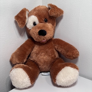 Könnte beinhalten: Ein brauner Plüschhund mit einem weißen Fleck über einem Auge, weißen Pfoten und Schlappohren. Das Stofftier hat eine schwarze Nase und eine rosa Zunge. Es sitzt auf einer weißen Oberfläche.